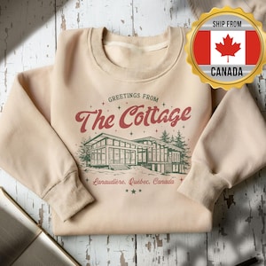 May include: A cream-coloured sweatshirt featuring the text "Greetings From The Cottage" in red, with a green illustration of a modern cottage. The text "Lanaudière, Québec, Canada" is below the cottage.
