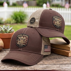 Puede incluir: Dos gorras de béisbol marrones con parte trasera de malla beige. Cada gorra tiene un parche en forma de escudo con un águila, el texto United States 250th Anniversary 1776-2026 y una pequeña bandera estadounidense en el lateral. El texto Breathable mesh back for all-day comfort es visible.