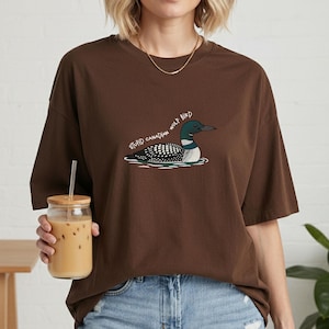 May include: A brown t-shirt with a graphic of a loon bird and the text "STUPID CANADIAN WOLF BIRD". The bird is illustrated in black, white, and teal, on water. The shirt has a relaxed, oversized fit.
