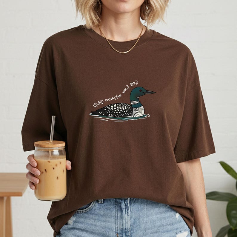 May include: A brown t-shirt with a graphic of a loon bird and the text "STUPID CANADIAN WOLF BIRD". The bird is illustrated in black, white, and teal, on water. The shirt has a relaxed, oversized fit.