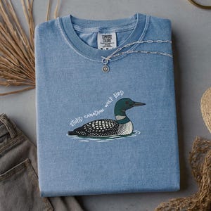 May include: A blue Comfort Colors t-shirt featuring a loon bird graphic and the text "STUPID CANADIAN WOLF BIRD". A silver necklace with a sun pendant is placed on the folded shirt. The shirt is a light blue colour.