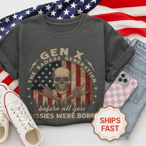 Peut inclure: T-shirt gris fonc&eacute; avec un motif repr&eacute;sentant un cr&acirc;ne, des mains de squelette et le texte "GEN X THE BADASS GENERATION before all you sissies were born" sur un motif de drapeau am&eacute;ricain. Le t-shirt est pr&eacute;sent&eacute; avec des baskets blanches et un jean bleu.