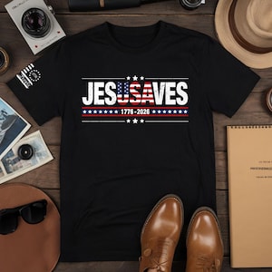 May include: A black short-sleeved t-shirt featuring the white text JESUSAVES with the letters USA stylized as an American flag. Below the text is the date 1776-2026 in white, flanked by horizontal lines and small white stars on a black background.