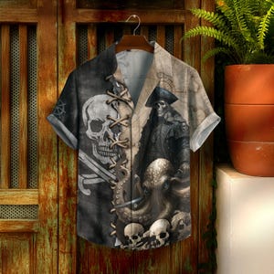 May include: Short-sleeved shirt with a pirate design. The shirt displays a skull, crossed swords, a pirate captain, and an octopus. The colour scheme includes black, grey, and tan.