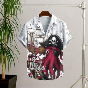 May include: Short-sleeved button-down shirt with a pirate-themed design. The shirt features a white background with illustrations of ships, and a large graphic of a red octopus wearing an eye patch and pirate hat. A skeleton and treasure chest are also featured.