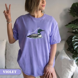 May include: A violet-coloured t-shirt with a graphic of a loon bird on water and the text "STUPID CANADIAN LOON BIRD". The shirt has short sleeves and is a solid colour. The word "VIOLET" is in a box at the bottom.