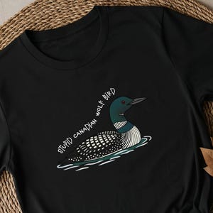 May include: Black t-shirt with a cartoon loon bird illustration. The bird has a black and white patterned body, a green head, and a black beak, floating on water. The text "STUPID CANADIAN WOLF BIRD" is written in white.