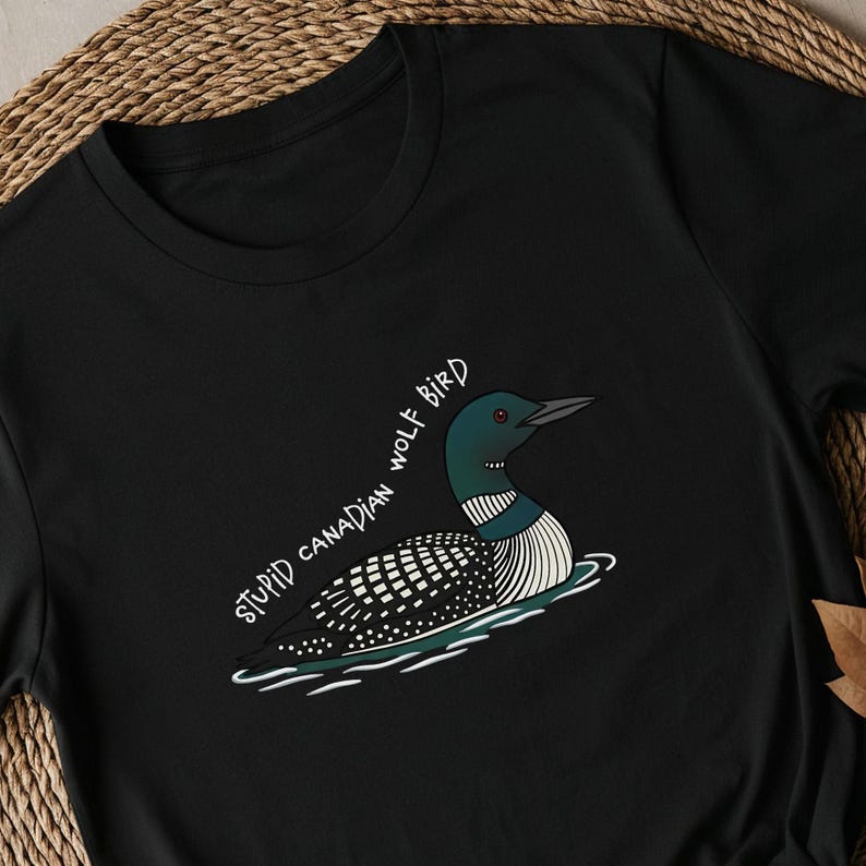 May include: Black t-shirt with a cartoon loon bird illustration. The bird has a black and white patterned body, a green head, and a black beak, floating on water. The text "STUPID CANADIAN WOLF BIRD" is written in white.