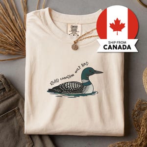 Peut inclure: T-shirt crème avec un dessin d'un huard dans l'eau, avec le texte "STUPID CANADIAN WOLF BIRD". Le t-shirt a un col rond et des manches courtes. Un collier doré avec un pendentif est visible.