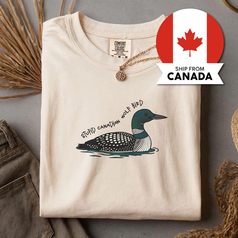 May include: A cream-coloured t-shirt featuring a graphic of a loon bird in water, with the text "STUPID CANADIAN WOLF BIRD". The shirt has a crew neck and short sleeves. A gold necklace with a pendant is visible.