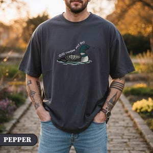 May include: Dark grey t-shirt featuring a loon illustration with the text "STUPID CANADIAN WOLF BIRD". The bird is depicted in water. The shirt has a relaxed fit and is made of a soft material.