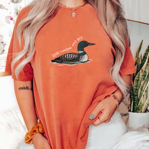 May include: Burnt orange t-shirt with a cartoon loon illustration and the text "STUPID CANADIAN WOLF BIRD". The loon is in black, white, and teal. The shirt is a casual wear item.