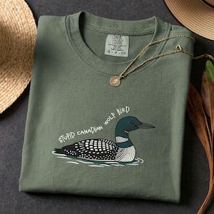 May include: An olive green t-shirt featuring a cartoon loon and the text "STUPID CANADIAN WOLF BIRD". The shirt has a crew neck and a small tag. A gold necklace with a pendant is draped across the shirt. The shirt is folded.