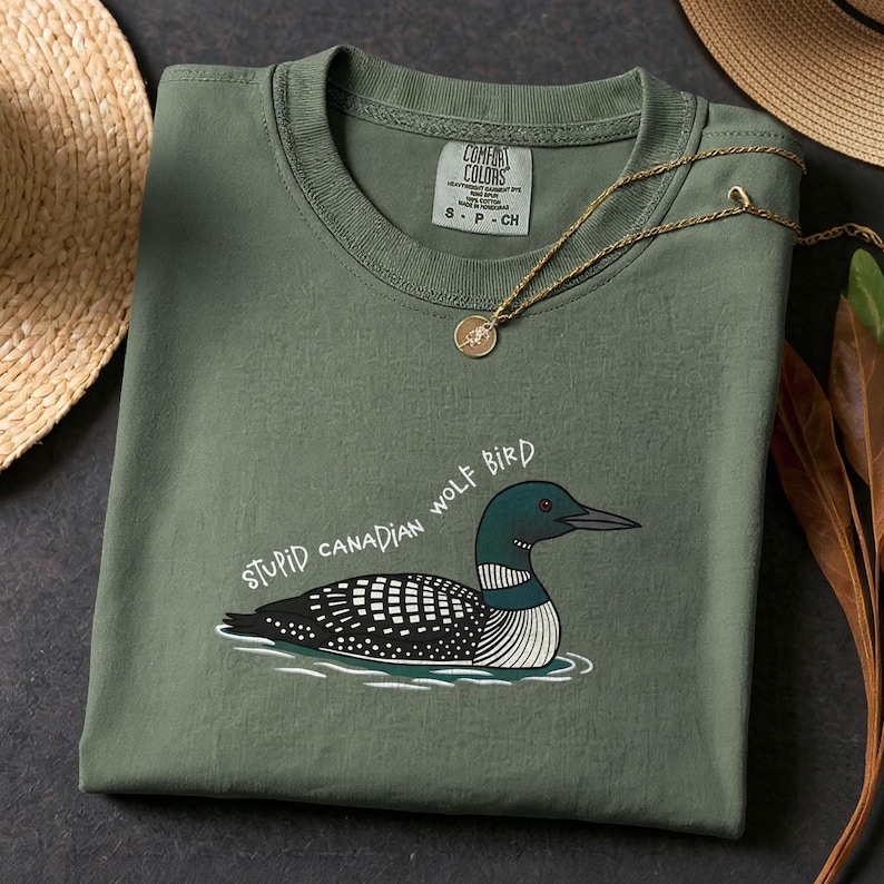 May include: An olive green t-shirt featuring a cartoon loon and the text "STUPID CANADIAN WOLF BIRD". The shirt has a crew neck and a small tag. A gold necklace with a pendant is draped across the shirt. The shirt is folded.