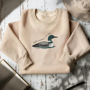 May include: A beige sweatshirt with a cartoon loon illustration and the text "STUPID CANADIAN WOLF BIRD". The loon has a black head, a white and black patterned body, and a green and white striped underbelly. The sweatshirt has a crew neck and ribbed cuffs.
