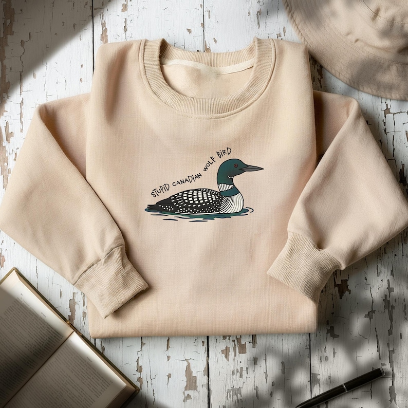 May include: A beige sweatshirt with a cartoon loon illustration and the text "STUPID CANADIAN WOLF BIRD". The loon has a black head, a white and black patterned body, and a green and white striped underbelly. The sweatshirt has a crew neck and ribbed cuffs.