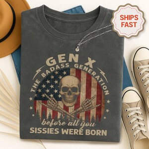 Peut inclure: T-shirt gris fonc&eacute; avec un motif de cr&acirc;ne, d'os crois&eacute;s et le texte "GEN X THE BADASS GENERATION before all you Sissies were born" sur un drapeau am&eacute;ricain. Un collier argent&eacute; et des baskets beiges sont &eacute;galement visibles.