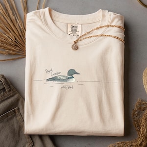 May include: A cream-coloured t-shirt featuring a loon illustration with the words "Stupid Canadian" and "Wolf bird". A gold necklace with a pendant is draped on the shirt. The t-shirt is folded.