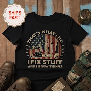 Puede incluir: Camiseta negra con un gráfico que presenta herramientas y la bandera estadounidense. El texto dice "THAT'S WHAT I DO, I FIX STUFF AND I KNOW THINGS". El diseño incluye un martillo, sierra, llave inglesa y otras herramientas.