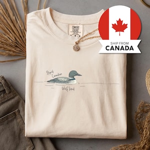 May include: A cream-coloured t-shirt with a cartoon illustration of a loon and the text "Stupid Canadian Wolf bird". The shirt has a crew neck and short sleeves. A gold necklace with a pendant is visible.