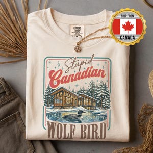 May include: A cream-coloured t-shirt with a graphic depicting a lake scene with a cabin, pine trees, and a loon. The text reads "Stupid Canadian" and "WOLF BIRD" in a retro style. A gold necklace is draped on the shirt.