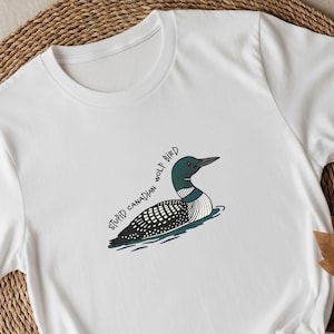 May include: White t-shirt with a cartoon loon bird illustration. The bird is shown in black, white, and green, floating on water. The text on the shirt says "STUPID CANADIAN WOLF BIRD".