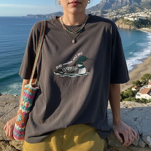 May include: Dark grey t-shirt with a cartoon loon and the text "STUPID CANADIAN WOLF BIRD." The outfit includes yellow trousers and a colourful crocheted bag. The background shows a scenic view of the ocean and mountains.