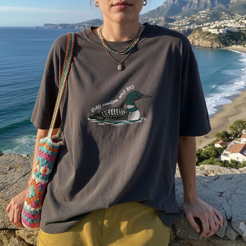 May include: Dark grey t-shirt with a cartoon loon and the text "STUPID CANADIAN WOLF BIRD." The outfit includes yellow trousers and a colourful crocheted bag. The background shows a scenic view of the ocean and mountains.