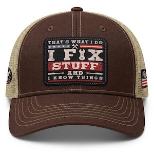 May include: A brown baseball cap with a tan mesh back and a rectangular patch on the front. The patch features the text "THAT'S WHAT I DO I FIX STUFF AND I KNOW THINGS" with a wrench icon and a stylized American flag design in red, white, and blue.