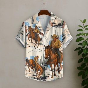 May include: Short-sleeved button-down shirt with a Western-themed print. The shirt features illustrations of cowboys on horseback, cacti, and desert landscapes in shades of brown, beige, and blue. The shirt is hanging on a wooden hanger.