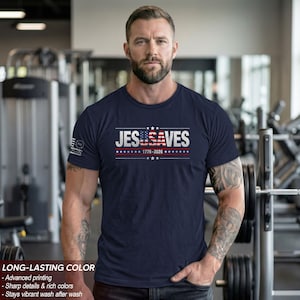 May include: A navy blue short-sleeve t-shirt featuring the white text JESUSA VES with the letters USA stylized as an American flag. Below the text is 1776-2026 with stars and horizontal lines. A small American flag emblem is on the right sleeve.