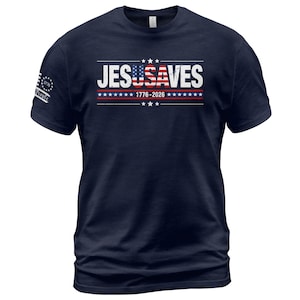 May include: A navy blue short-sleeve t-shirt featuring the white text JESUSAVES with the letters USA stylized as an American flag. Below the text is a horizontal line of white stars and the numbers 1776-2026. A small 1776 logo is on the sleeve.