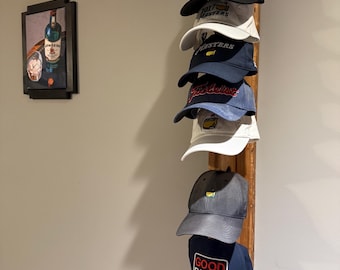 Puede incluir: Un estante de madera para sombreros que muestra siete gorras de béisbol de varios colores, incluyendo negro, blanco y azul marino, con texto y logotipos. El estante está montado en una pared de color claro, con un cuadro enmarcado a la izquierda.