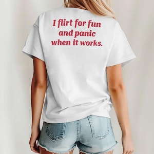 Könnte beinhalten: Weißes T-Shirt mit dem Text "I flirt for fun and panic when it works" in roter Schrift. Das Shirt hat kurze Ärmel und wird mit Jeansshorts getragen. Das Bild zeigt den Rücken einer Person.