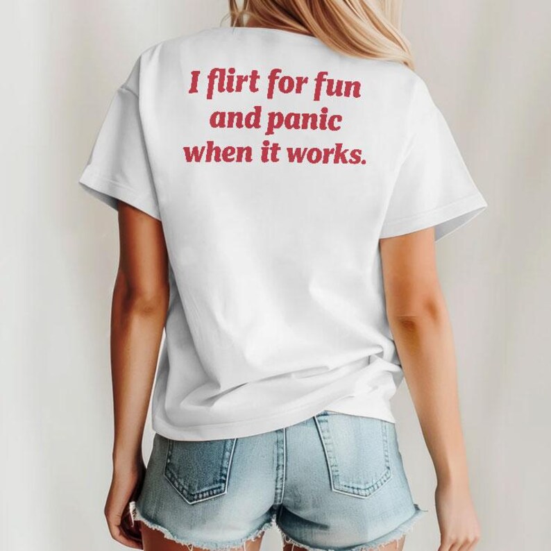 May include: White t-shirt with the text "I flirt for fun and panic when it works" in red lettering. The shirt is short-sleeved and worn with denim shorts. The image shows the back of a person.