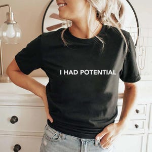 Könnte beinhalten: Schwarzes T-Shirt mit Rundhalsausschnitt und dem weißen Text "I HAD POTENTIAL". Das Shirt besteht aus einem weichen Material und eignet sich für Freizeitkleidung. Das Shirt wird mit einer blauen Jeans kombiniert.