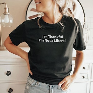 May include: Black crew neck t-shirt with the white text "I'm Thankful I'm Not a Liberal". The shirt is made of a soft material and is suitable for casual wear. The shirt is paired with blue jeans.