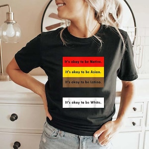 Puede incluir: Camiseta negra con un diseño rectangular que incluye el texto "It's okay to be Native", "It's okay to be Asian", "It's okay to be Latino" y "It's okay to be White". El texto es blanco y sin serifa.