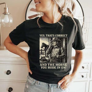 Puede incluir: Camiseta negra con una ilustración retro de un vaquero hablando por teléfono, un caballo y el texto "YES, THAT'S CORRECT AND THE HORSE YOU RODE IN ON". El diseño es de estilo vintage.