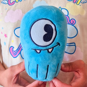May include: A plush toy shaped like a rounded blue monster with a single large embroidered eye, a curved mouth, and two small white teeth. The bottom of the toy features four vertical stitched lines, and the surface is made of soft blue fabric.