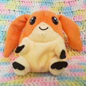 May include: A plush toy featuring a bright orange head with long, drooping ears, large black eyes, and a small stitched smile. The body is a pale cream color with four short limbs, each ending in small, rounded black paws or feet.