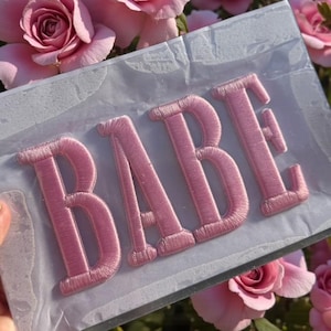 Puede incluir: Un parche bordado rosa con la palabra "BABE" en mayúsculas. El parche está sobre un respaldo transparente, con un fondo de rosas rosas.