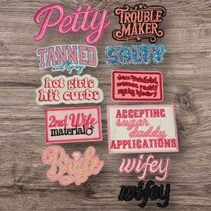 Peut inclure: Plusieurs écussons brodés avec des phrases en rose, noir et bleu. Les phrases incluent "Petty", "Trouble Maker", "Tanned and tipsy", "Salty", "Hot girls hit curbs", "2nd Wife material", "Bride" et "Wifey".