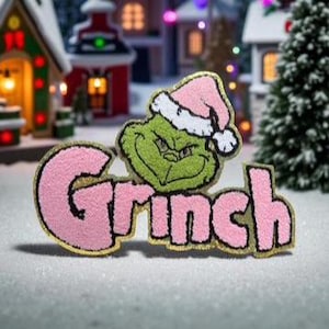 Puede incluir: Un parche bordado en rosa y dorado con la palabra "Grinch" en una fuente lúdica. El parche incluye una imagen del Grinch con un gorro de Papá Noel rosa. El parche está sobre un fondo nevado con edificios de un pueblo navideño en miniatura.