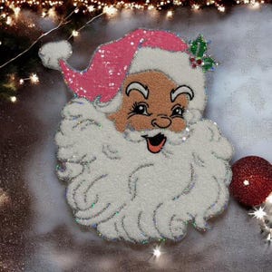 May include: A festive Santa Claus decoration with a pink sequined hat and a fluffy white beard. The Santa face has a rosy complexion and a smiling expression. A red ornament is visible on the right side of the image. The decoration is outlined with silver glitter.