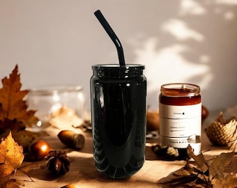 Puede incluir: Un vaso negro con una pajita negra. El vaso está lleno de un líquido oscuro. A la derecha hay una pequeña vela en un tarro de cristal. La escena incluye hojas de otoño y bellotas.