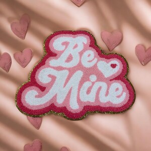 May include: A pink and white rug with the words "Be Mine" in a cursive font. The rug has a gold glitter border and is surrounded by small pink heart-shaped decorations. The rug is on a pink background.