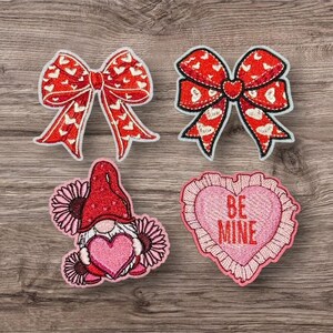 May include: Four embroidered patches on a wood grain surface. Two red bows with white heart patterns, a gnome holding a pink heart, and a pink heart with "BE MINE" text.