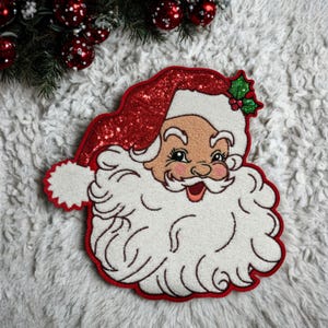 Puede incluir: Un parche festivo con la cara de Papá Noel, con un gorro rojo con lentejuelas y barba blanca. El parche tiene un borde rojo y una cara sonriente con mejillas rosadas. Una ramita de acebo está en el gorro. Ideal para manualidades y decoraciones navideñas.