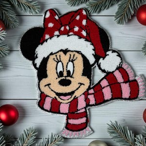 Peut inclure: Décoration de Noël Minnie Mouse. Minnie porte un nœud rouge à pois blancs, un bonnet de Père Noël bordé de blanc et une écharpe rayée rouge et rose. La décoration est entourée de branches de pin et de boules rouges et argentées.